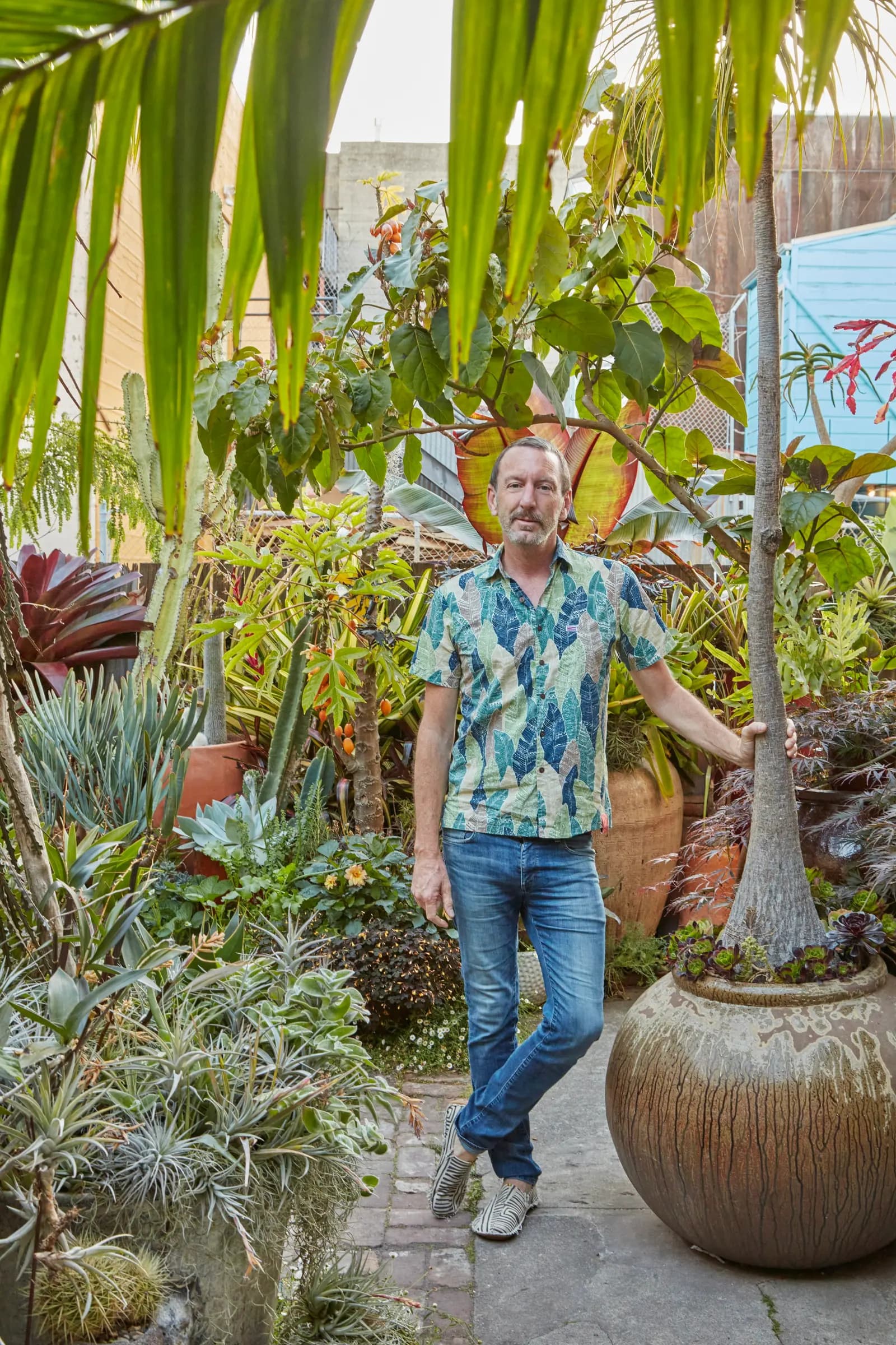 Un hombre con camisa colorida de estampado de hojas y jeans, de pie en un jardín exuberante rodeado de plantas en macetas y vegetación.