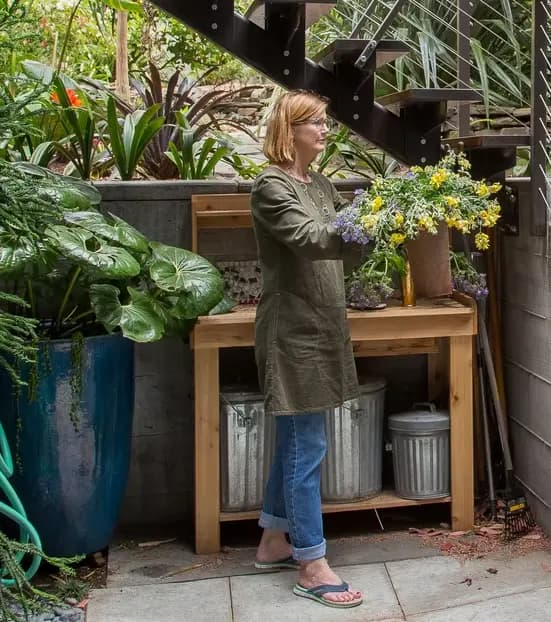Una mujer rubia con lentes en un jardín, con un ramo de flores amarillas y moradas, junto a una mesa de madera rodeada de plantas grandes y artículos de jardinería.