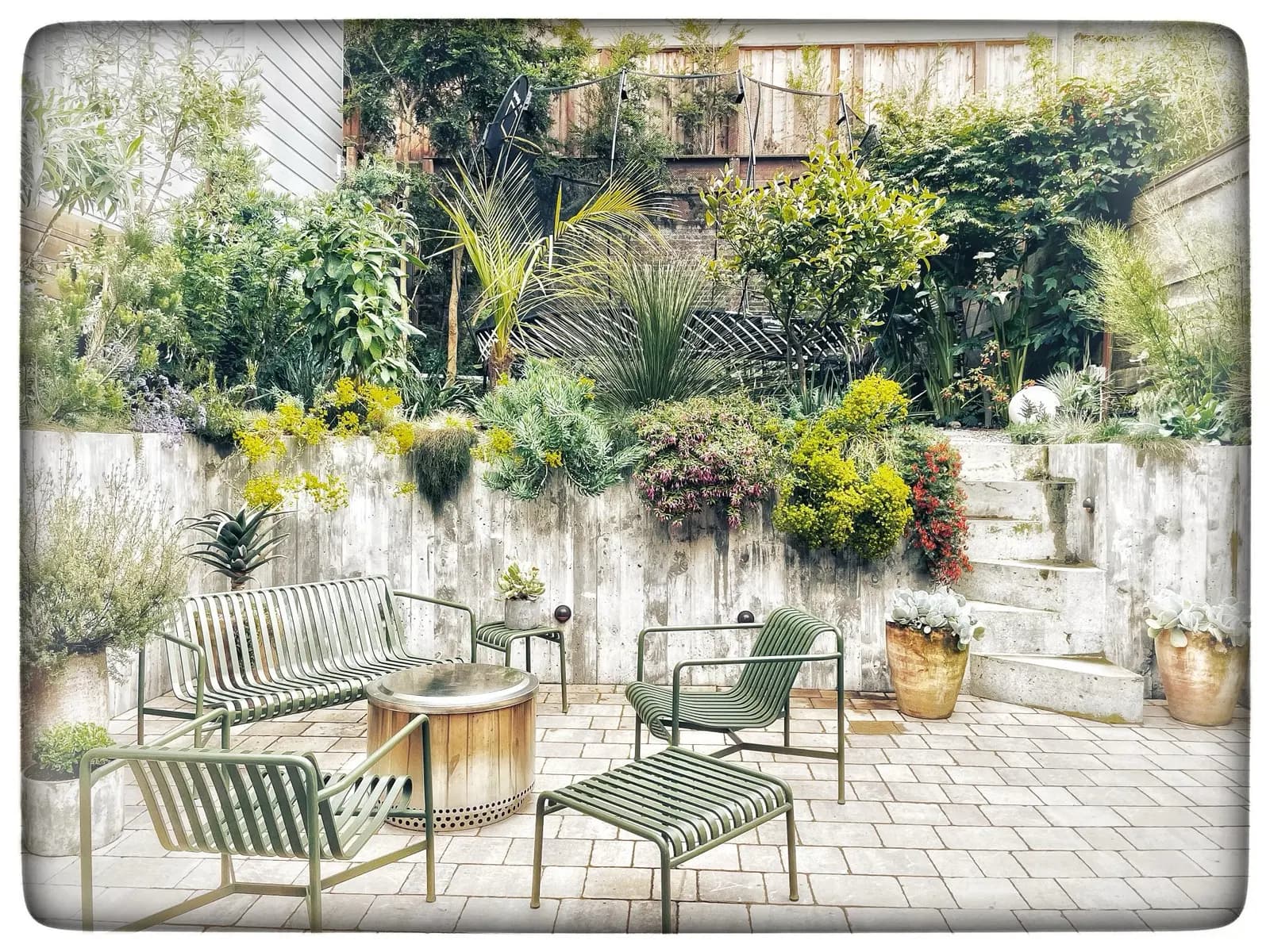 Un patio acogedor con árboles, plantas y flores sobre concreto y ladrillo, con un fogón redondo rodeado de sillas de metal con cojines a rayas.