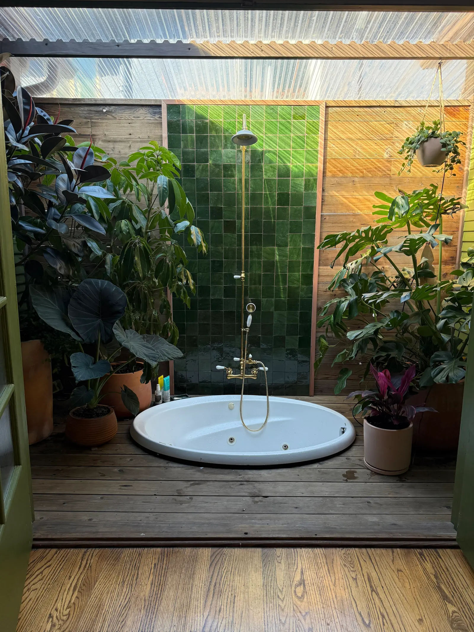 A bathroom greenhouse with an outdoor bath tub and shower space that has green tile walls, wooden floor, and a ceiling made of transparent corrugated material. The outdoor space contains potted plants. Designed with Terri Loenthal by Clarke De Mornay