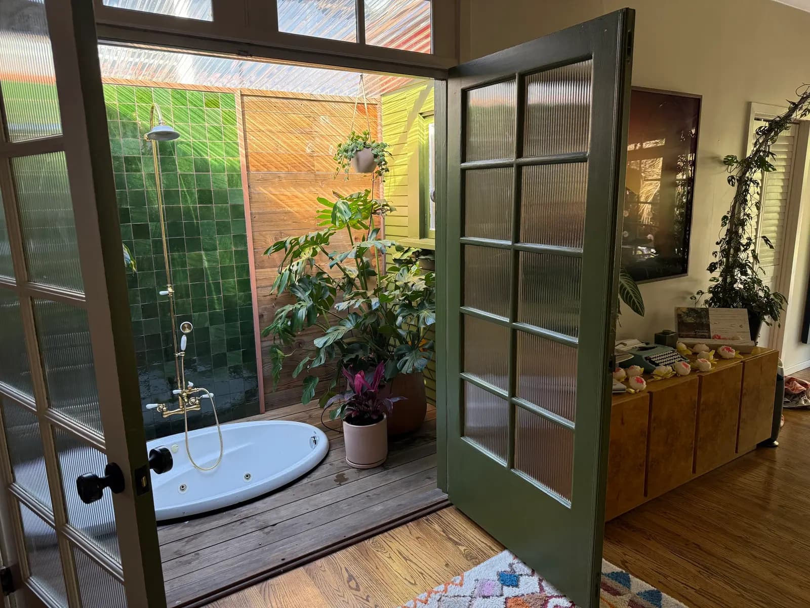 Un baño tipo invernadero con tina y regadera al aire libre, paredes de azulejo verde, piso de madera y techo de lámina corrugada transparente. El espacio exterior tiene plantas en macetas. Diseñado por Clarke De Mornay con Terri Loenthal