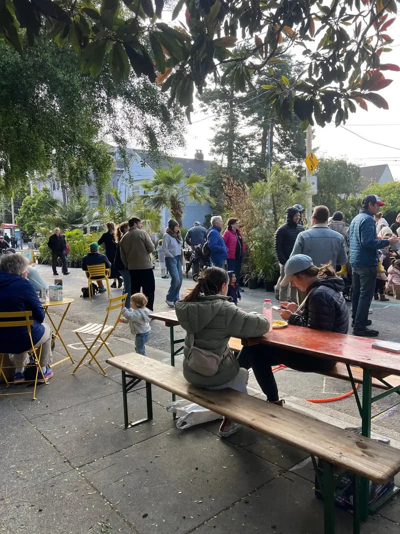 Personas reunidas en la plaza pop-up de Cole Valley creada por Flora Grubb. Algunas están sentadas a las mesas y otras hacen fila o platican. Hay niños jugando y plantas en macetas cerca.