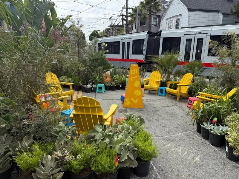 Una plaza temporal en Cole Valley hecha por Flora Grubb Gardens, con un área de asientos al aire libre llena de color, sillas amarillas, mesitas y plantas en macetas, y un tren que pasa al fondo.