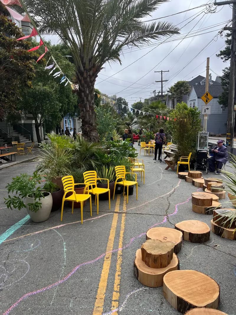 La plaza de Cole Valley de Flora Grubb Gardens, con sillas amarillas, plantas en macetas, troncos, dibujos de gis y personas caminando y sentadas, bajo un cielo nublado.