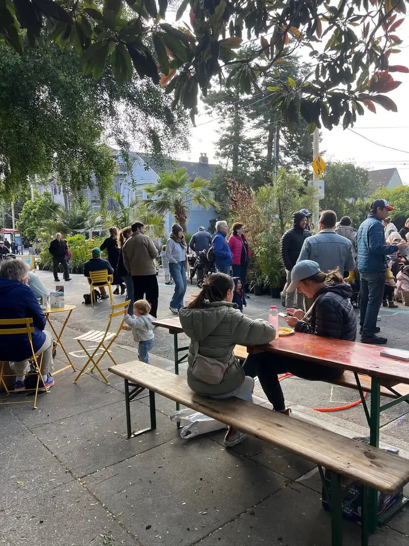 Personas reunidas en la plaza pop-up de Cole Valley creada por Flora Grubb. Algunas están sentadas a las mesas y otras hacen fila o platican. Hay niños jugando y plantas en macetas cerca.