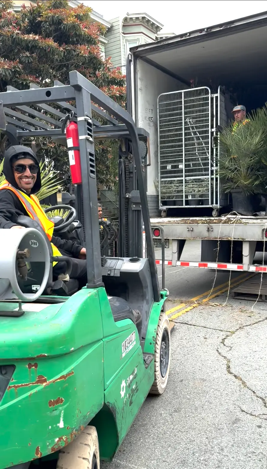 Un operador de montacargas con chaleco de seguridad y lentes de sol sonríe mientras trabaja al aire libre junto a un camión lleno de plantas en macetas y vegetación, con dos personas al fondo.