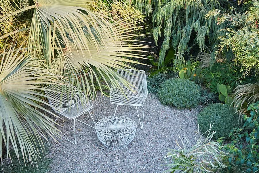 El jardín de la casa de Flora Grubb con dos sillas blancas de alambre y una mesita de alambre a juego, rodeadas de exuberantes plantas tropicales y arbustos, sobre un piso de grava.