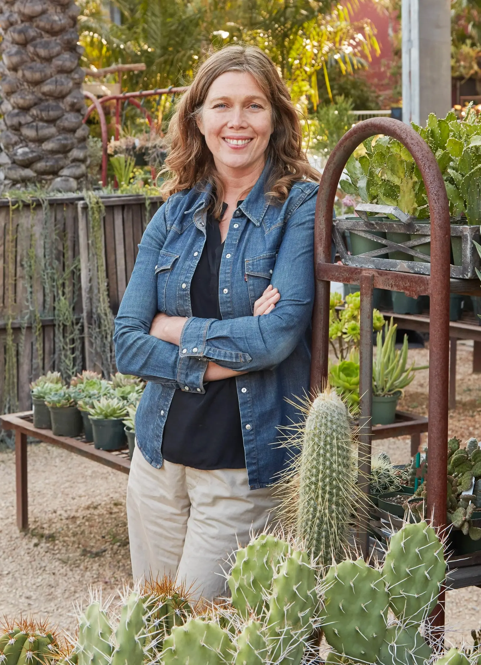 Flora Grubb, fundadora y dueña de Flora Grubb Gardens, sonriendo con los brazos cruzados en Flora Grubb Gardens San Francisco, con una chamarra de mezclilla sobre una camisa negra.