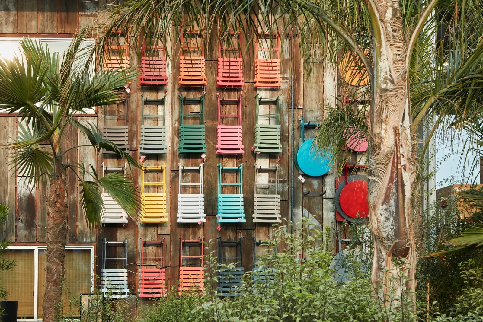 Muebles Fermob: sillas plegables de colores y mesitas plegables exhibidas en una pared de madera, rodeadas de árboles y plantas.