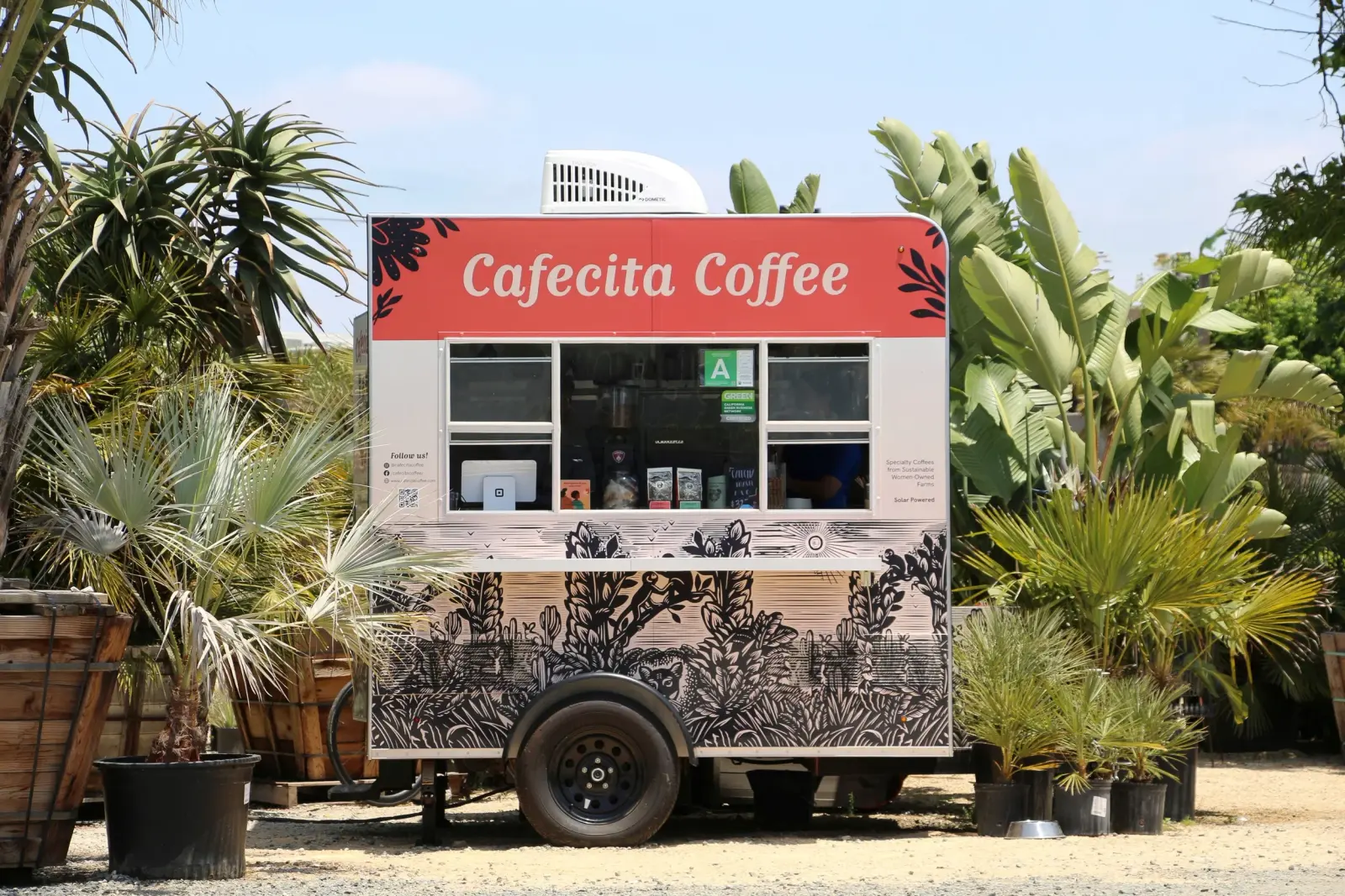 El tráiler de Cafecita Coffee rodeado de plantas en macetas y follaje tropical bajo un cielo azul en Flora Grubb Gardens