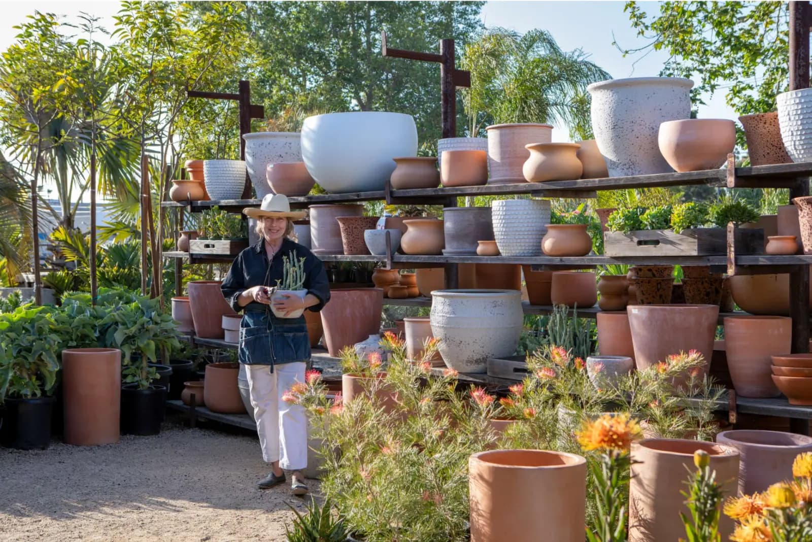 A woman in a black shirt and white apron holding a potted plant outdoors at a garden center. Behind her are shelves filled with various large, empty ceramic pots. She is wearing a wide-brimmed hat, and there are green trees and colorful flowering plants around her.
